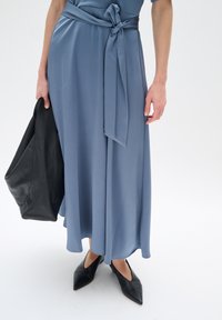 Langes, blaues Kleid mit strukturiertem Finish und gebundenem Taillengürtel. Kombiniert mit spitzen schwarzen Schuhen und einer lässigen schwarzen Ledertasche.