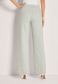 Vrouw in lichtgrijze, hoge taille met wijde pijpen en subtiel patroon, gecombineerd met witte hoge hakken, staand tegen een bleekgekleurde muur.