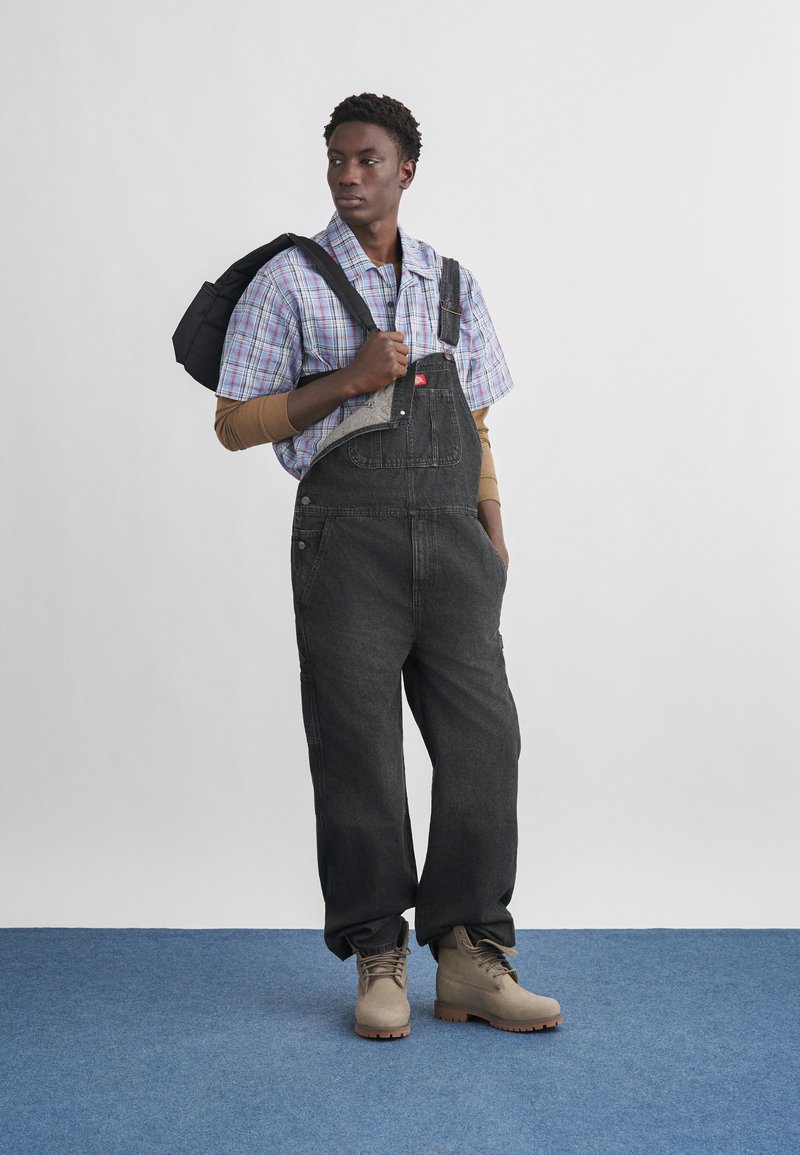 Giovane uomo che indossa una salopette di denim nera, una camicia a maniche corte a quadri sopra una maglia a maniche lunghe color sabbia, stivali beige e porta una borsa nera su una spalla.