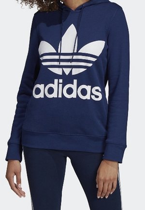 Personne portant un sweat à capuche Adidas bleu marine avec logo blanc et un pantalon bleu marine assorti avec des bandes blanches sur les côtés.