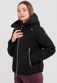 Schwarze Pufferjacke mit Kapuze, ausgestattet mit metallischen Reißverschlüssen, Seitentaschen und einem strukturierten Muster auf dem Stoff. Silberne Hardware-Details.