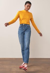Gul langærmet bluse parret med høj talje blå jeans og røde sneakers. Baggrunden er neutral med trægulv.