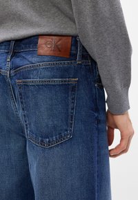 Jeans in denim blu scuro con una patch in pelle di Calvin Klein sul retro, dotati di tipiche tasche posteriori e dettagli di cucitura a contrasto.