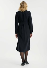 Vestido negro de manga larga con cintura entallada y dobladillo acampanado, confeccionado en tela suave. Presenta un cuello y puños abotonados. Un par de tacones negros.
