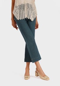 Top in cotone beige all'uncinetto, jeans a gamba larga blu scuro e scarpe con tacco a blocco beige lucide con dettaglio della fibbia quadrata. Texture liscia in tutto.