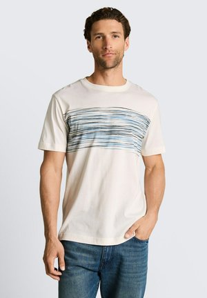 Mężczyzna w kremowej koszulce na krótki rękaw z poziomym wzorem w niebieskie i czarne paski oraz w niebieskich dżinsach, stojący z jedną ręką w kieszeni.