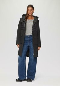 Schwarzer Mantel mit gestepptem, glänzendem Vorderteil, Kapuze und Taschen. Kombiniert mit einem gestreiften Oberteil und weit geschnittenen blauen Jeans. Schwarze klobige Stiefel.