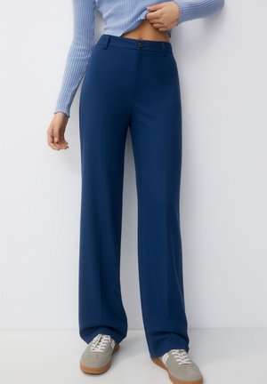 Vrouw die marineblauwe wijde broek met hoge taille, lichtblauwe geribde trui en grijze sneakers draagt, staand tegen een witte achtergrond.