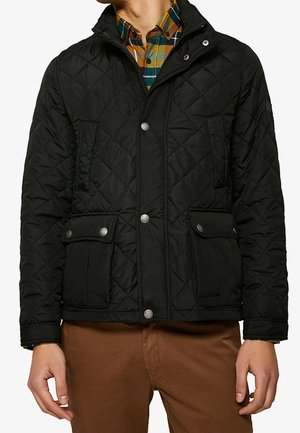 Giubbotto trapuntato nero con collo alto, chiusure a pressione e due tasche frontali. Indossato sopra una camicia a quadri multicolore.