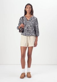 Svart og hvit mønstrete bluse med V-hals, pufferm; beige shorts; brune sko med teksturerte såler; holder en liten mørk veske.