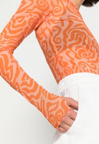 Oranje top met lange mouwen en abstracte krullen, gemaakt van doorschijnende stof. Witte broek met een zak zichtbaar aan de rechterkant.