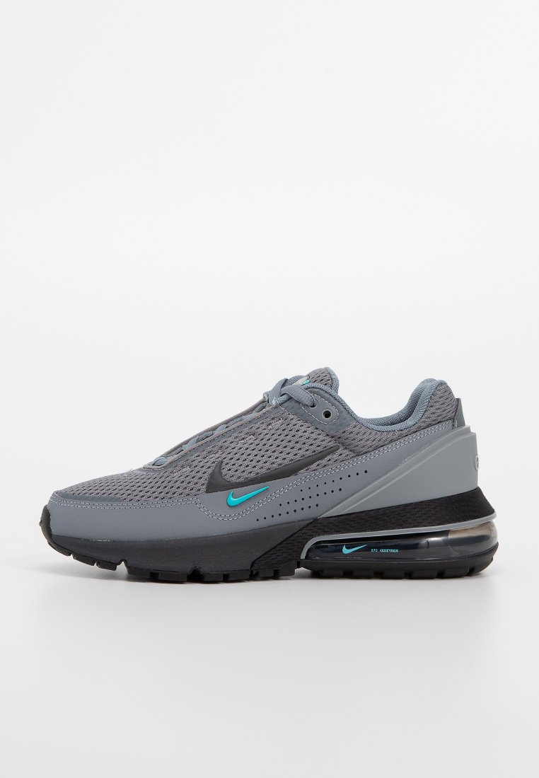 Zapatilla deportiva gris con parte superior de malla, suela de goma negra, acentos en color teal y una unidad de amortiguación visible. Punta redondeada y diseño de cordones.