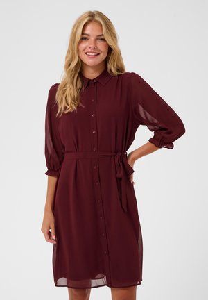 Femme souriante aux longs cheveux blonds portant une robe bordeaux boutonnée avec des manches trois-quarts et une ceinture nouée à la taille sur fond blanc.