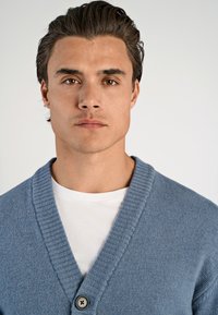 Ljusblå stickad cardigan med en ribbad sjalkrage och en enda svart knapp, bärs över en vit t-shirt. Slät textur och avslappnad design.