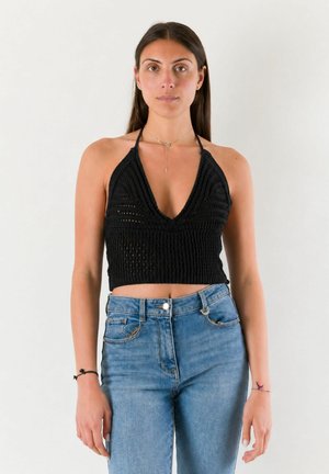 Donna che indossa un top halter nero all'uncinetto e jeans blu a vita alta, in piedi davanti a uno sfondo bianco semplice.