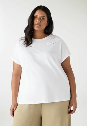 Donna con capelli lunghi e scuri, indossa una maglietta bianca a maniche corte con ritagli floreali ricamati e pantaloni beige a gamba larga, in piedi su sfondo bianco.