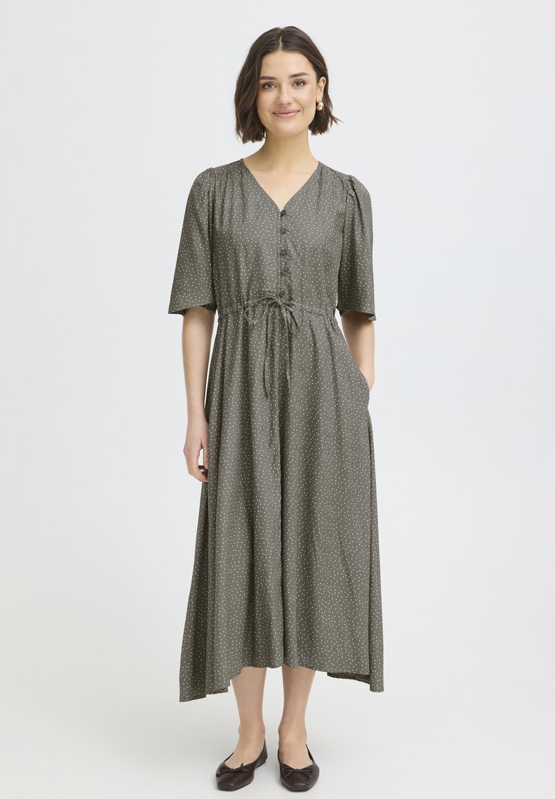 Une femme souriante porte une robe midi grise à pois avec des boutons sur le devant, des manches mi-longues et des ballerines marron, sur un fond blanc.