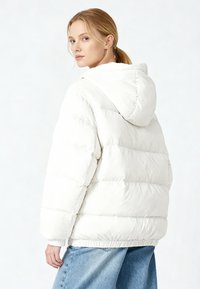 Doudoune blanche avec capuche, comprenant des sections matelassées et des poignets élastiques. Portée avec un jean denim bleu, vue de dos.