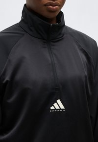 Close-up of a person wearing a black quarter-zip pullover with a high collar and a white Adidas Basketball logo on the chest.