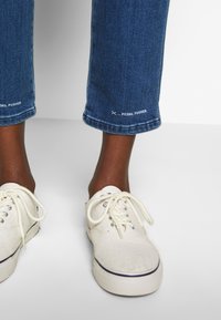 Jambes à la peau foncée portant un jean bleu court marqué "PEDAL PUSHER" au-dessus de baskets blanches en toile avec lacets.