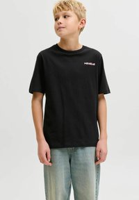 Blond jongetje met een zwart T-shirt met korte mouwen en kleine kleurrijke tekst op de borst, gecombineerd met lichtblauwe spijkerbroek, staand tegen een effen achtergrond.