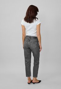 Žena s hnedými vlasmi po pleciach, oblečená v bielom tričku s krátkym rukávom, čiernych nohaviciach s vysokým pásom a čiernych nízkych podpätkoch, stojí.