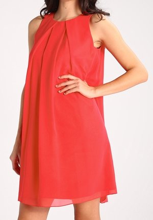 Kvinde iført en ærmeløs, knælængde rød kjole med plisseret forside, poserer med den ene hånd på hoften mod en ensfarvet baggrund.