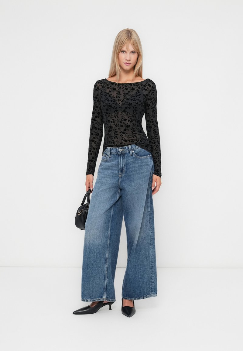 Schwarzes Langarmoberteil mit floralem Muster, kombiniert mit hellblauen Weitbeinhosen und schwarzen spitzen Schuhen. Kleine schwarze Handtasche.