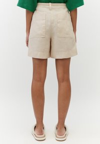 Shorts in lino beige con due tasche posteriori, orli risvoltati e vita senza cuciture, abbinati a scarpe slip-on neutre.