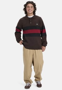 Braun gestrickter Poloshirt mit roten und schwarzen horizontalen Streifen, drei Knöpfen am Kragen und entspannten beigen Hosen.