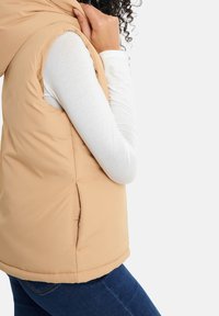 Beige Puffer-Weste mit Kapuze, ärmellosem Design, seitlichen Schlitzen und glatter Oberflächenstruktur, getragen über einem weißen Langarmshirt.