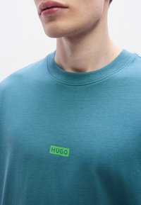 Μπλε-πράσινο βαμβακερό t-shirt με λαιμόκοψη και μικρό πράσινο λογότυπο "HUGO" στο στήθος. Μαλακή υφή χωρίς ορατά σχέδια ή διακοσμητικά στοιχεία.