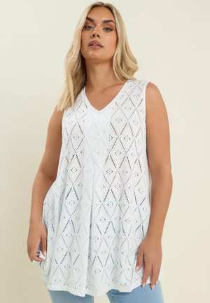 Woman with long blonde hair wearing a sleeveless white eyelet-patterned V-neck tunic over light blue jeans, standing against a plain neutral background.