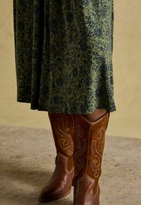 Długa zielona sukienka w ciemnoniebieski kwiatowy wzór, w zestawieniu z brązowymi zamszowymi kowbojkami ozdobionymi haftowanymi zwojami i zwężającym się krojem.