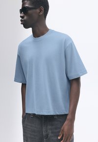 T-shirt oversized blu chiaro con maniche corte, scollatura rotonda e texture morbida. Indossato con jeans grigi scuri; occhiali da sole neri completano il look.