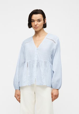 Νεαρή γυναίκα με κοντά σκουρόχρωμα μαλλιά, που φοράει μια ανοιχτομπλε ριγέ μακρυμάνικη μπλούζα και άσπρο παντελόνι, στέκεται μπροστά σε ένα απλό φόντο.