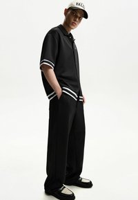 Polo nero a maniche corte con strisce bianche, pantaloni neri a gamba larga e scarpe nere con dettagli beige texturizzati, abbinati a un cappellino bianco.