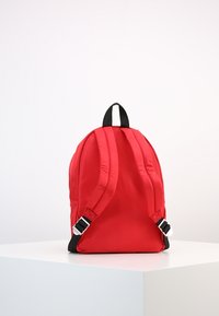 Röd tygryggsäck med vadderade axelremmar, svarta detaljer och silverfärgade detaljer. Rundad toppdesign och slät yta. Justerbara spännen.