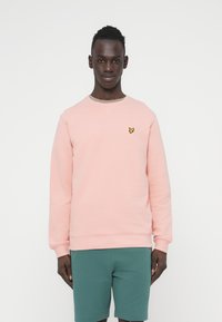 Φούξια ροζ φούτερ από βαμβάκι, με στρογγυλή λαιμόκοψη και ριμπ μανσέτες, με ένα μικρό κίτρινο λογότυπο πεταλούδας στο στήθος. Φοριέται με πράσινα σορτς.