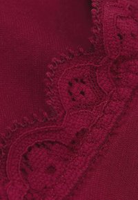 Tela burdeos con bordes de encaje intrincado que presenta bordes en forma de concha y pequeños patrones recortados, resaltando una superficie texturizada.