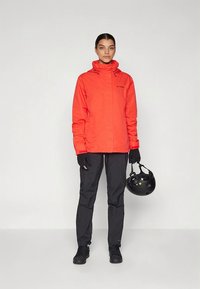 Svetlo oranžna vodoodporna jakna s kapuco, v kombinaciji s črnimi hlačami in rokavicami. Drži črno čelado. Enostavna, funkcionalna zasnova.