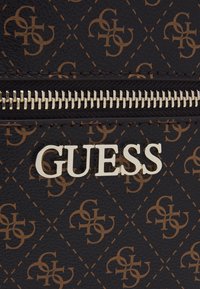 Lähikuva ruskeasta kuviollisesta Guess-laukusta, jossa on hopeinen vetoketju ja metallinen Guess-logo edessä.