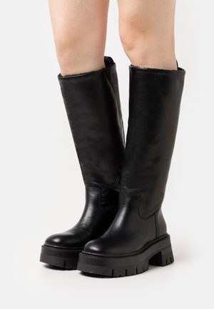 Botas de cuero negro hasta la rodilla con una suela de goma gruesa, superficie lisa, punta redonda y un forro interior suave visible en la parte superior.