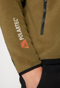Olivengrøn fleecejakke med sort kant, der har en lynlås krave og hvidt Polartec-logo med rød accent på ærmet.