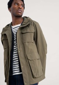 Veste à capuche vert olive avec quatre poches frontales, fermeture éclair et boutons-pression. Superposée sur un cardigan noir à boutons et une chemise rayée.