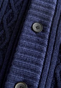 Cardigan a maglia blu navy con texture a coste verticali e un dettaglio con bottone nero. Il tessuto presenta una finitura morbida e strutturata.