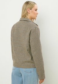 Giacca a motivi houndstooth marrone e beige con colletto, maniche lunghe e taglio dritto. Indossata sopra jeans azzurri chiari.
