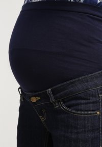 Tmavě modré těhotenské džíny se strečovým černým pasem, zdobené zlatým prošitím a předním zapínáním na knoflík. Hladký povrch látky.