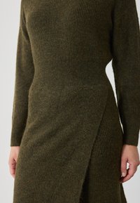 Olívazöld kötött ruha texturált mintával, hosszú ujjakkal és derékra csavarható fazonnal, mely puha, kényelmes anyagból készült.