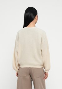 Suéter bege claro com um tricot texturizado, apresentando ombros caídos, mangas longas e um ajuste solto. Combinado com calças bege.
